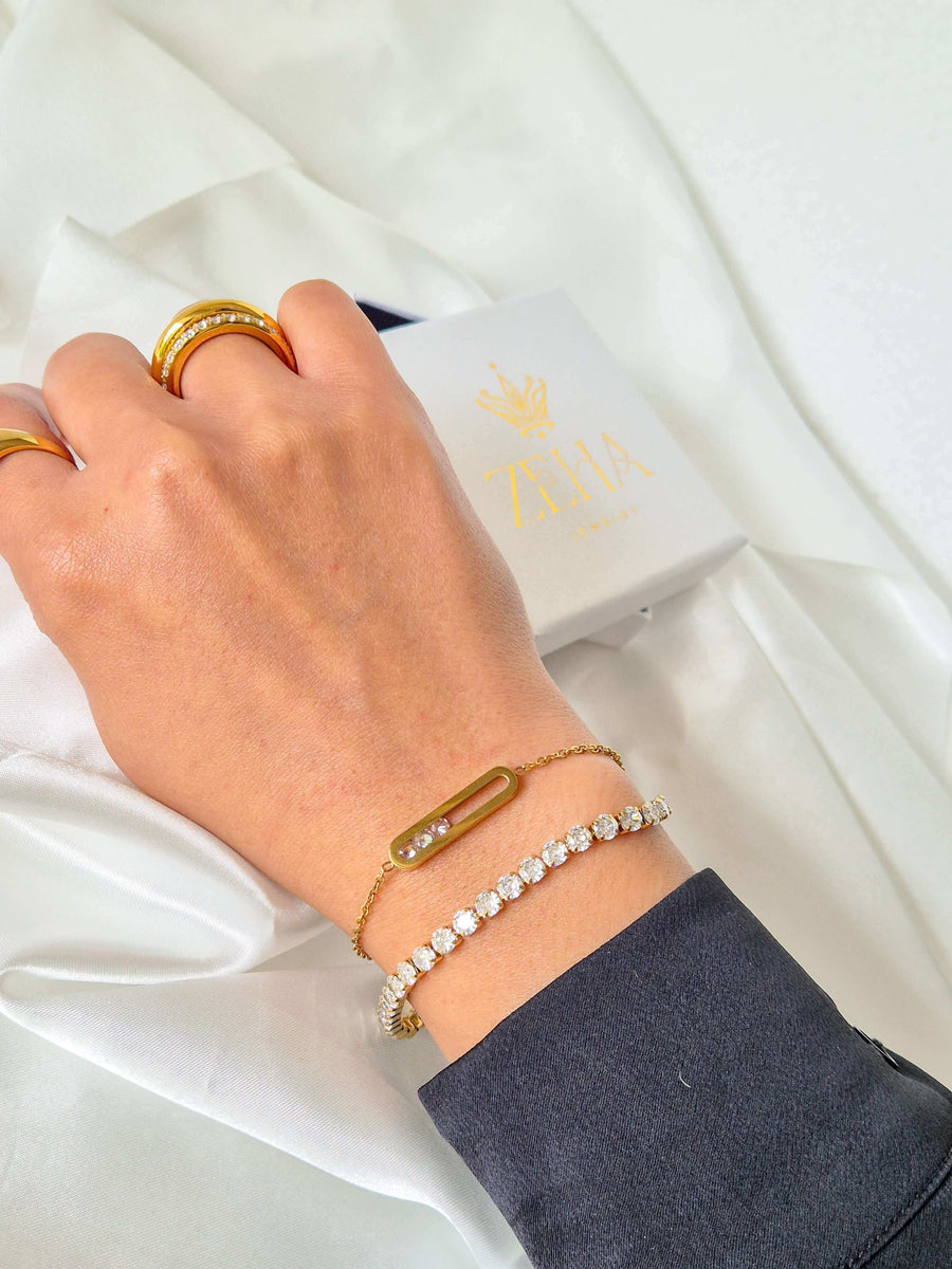 Tennisarmband mit runden klaren Steinen und goldfarbenes Kettenarmband mit länglichem Anhänger, am Handgelenk getragen