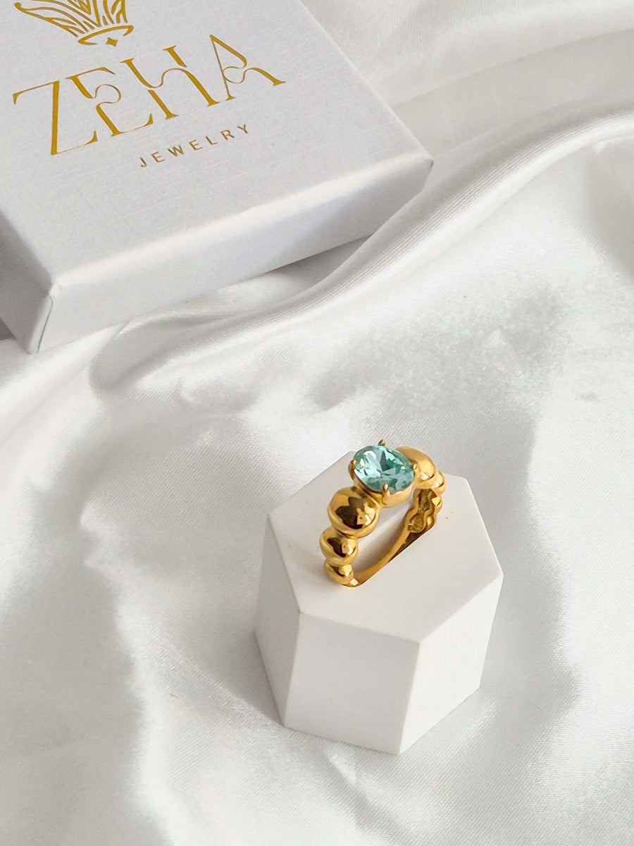 Goldfarbener Ring mit ovalem aqua-blauem Zirkonia auf weißem Schmuckständer