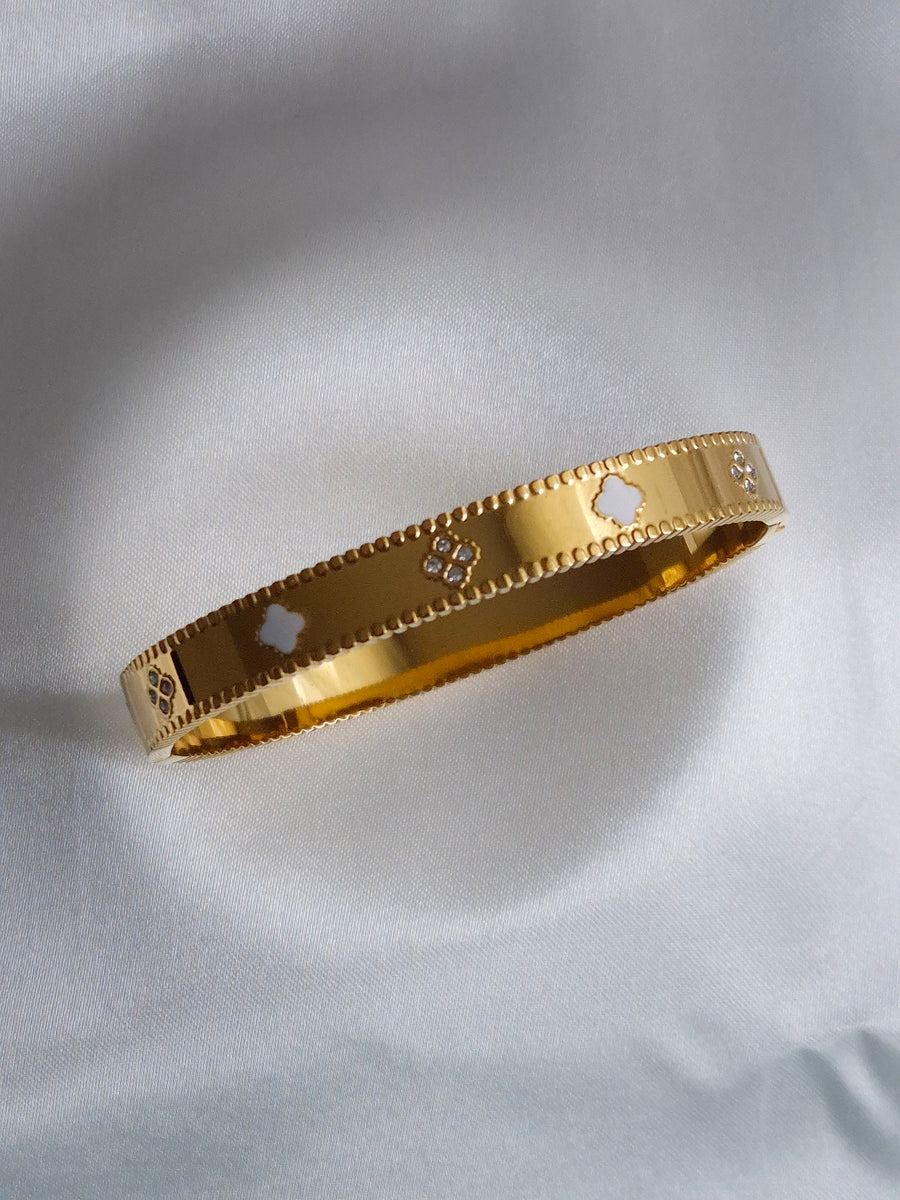 Goldfarbener Kleeblatt-Armreif mit weißen Motiven, klaren Steinen und gerillten Rändern auf weißem Stoff